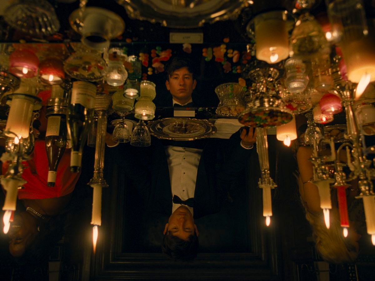 Oliver (Barry Keoghan), in black tie dress, sits at what appears to be an fancy table covered in candles of all descriptions, reflecting his face back at him —&nbsp;except the more you look, the more it’s clear that the reflection is in a different position, standing with its eyes lowered. From the movie Saltburn