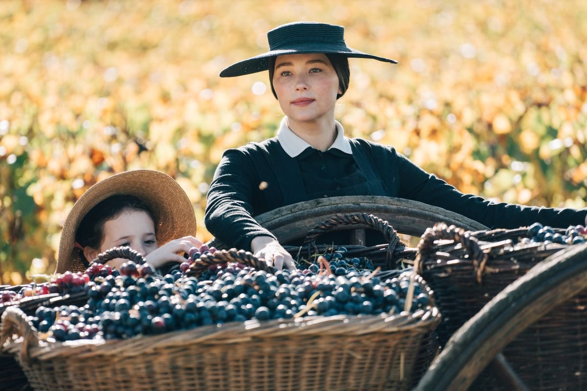 A woman in a black dress and hat standing next to a basket of grapes in front of a field of corn in Widow Clicquot.