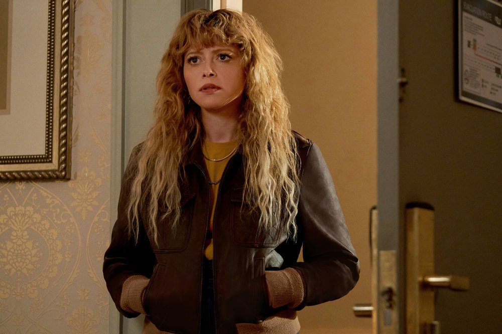 Natasha Lyonne as Charlie Cale, wearing a leather jacket in a yellow-hued hotel room, standing in the door frame, in Poker Face